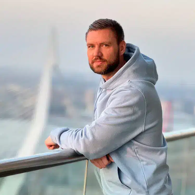Man in grijze hoodie leunt op glazen balkon met uitzicht over stad aan het water.